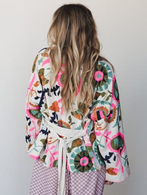 A back-view shot of a heavily embroidered kimono-style jacket in cream with bold floral and botanical motifs in hot pink, olive, teal, rust, and navy — tied at the waist and layered over a lavender geometric-print bottom.