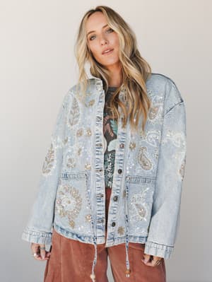An oversized acid-wash denim jacket covered in intricate paisley embroidery and iridescent sequin detailing, layered over a graphic tee and rust wide-leg pants — the kind of statement piece that looks like it has a really good story behind it.