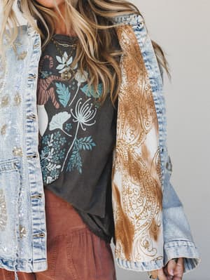 A close-cropped lifestyle shot of a layered boho outfit — a botanical graphic tee and rust bottoms peeking out beneath an embellished denim jacket with a striking paisley-printed lining, styled with gold layered necklaces for that effortless, collect