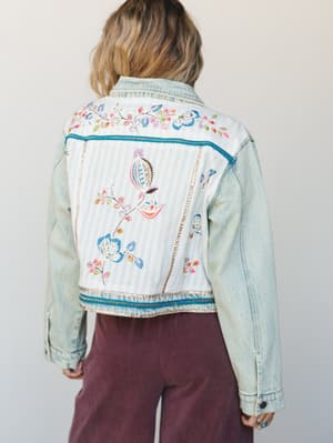 A back-facing model shows off a light wash denim jacket with a patchwork-style back panel — soft white stripe fabric embroidered with colorful botanical florals, finished with teal ribbon and metallic trim details, paired with dusty mauve wide-leg pa