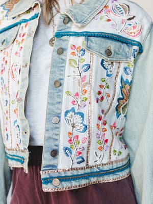A close-up of a light wash denim jacket covered in dense floral embroidery, sequin trim, and teal pom-pom edging — layered over a white tee and worn with a dusty mauve bottom, the whole thing feels like someone turned their favorite quilt into outerw