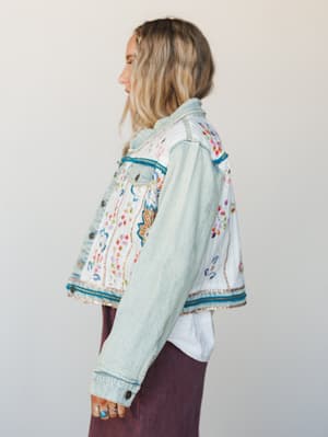 A light-wash denim jacket covered in colorful floral embroidery and beaded trim details, styled over a white tee and plum skirt — the kind of jacket that does all the talking.