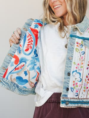 A close-up lifestyle shot showing the inside and outside of an embroidered denim jacket — the printed floral lining is held open to reveal intricate multicolor embroidery along the front placket, layered over a white tee and deep plum bottoms.