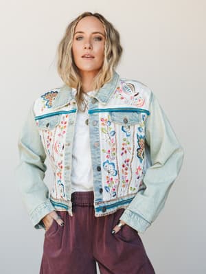 A relaxed, expressive denim jacket covered in colorful floral embroidery and teal trim details, layered over a white tee and paired with dusty plum wide-leg pants — the kind of piece that does all the talking.