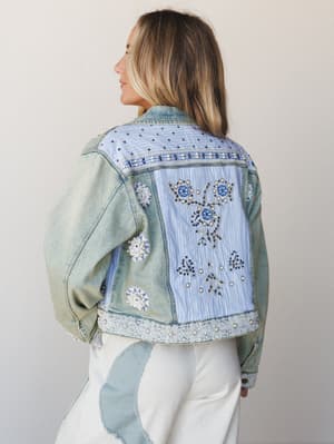A back-facing model shows off an embellished denim jacket with a patchwork striped panel, pearl and bead detailing, lace trim at the hem, and floral appliqués — the kind of jacket that tells a story before you say a word.