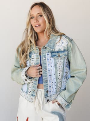 A light-wash denim jacket covered in lace trim, pearl beading, floral appliqué, and blue embroidery — the kind of jacket that tells a story before you even open your mouth — styled over a blue crop top and cream drawstring pants in a clean studio set