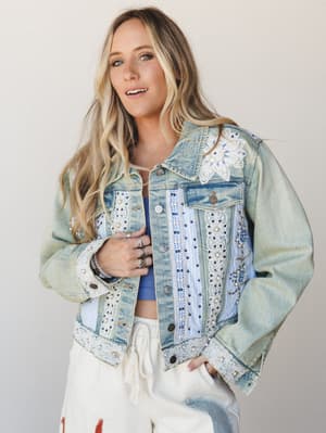A light-wash denim jacket covered in lace trim, pearl beading, floral appliqué, and blue embroidery — the kind of jacket that tells a story before you even open your mouth — styled over a blue crop top and cream drawstring pants in a clean studio set
