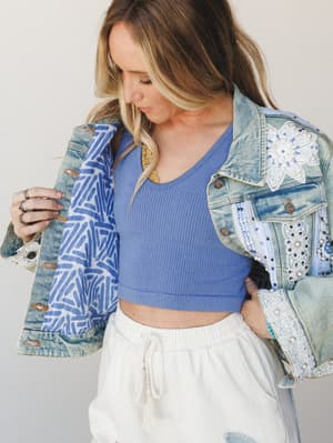 A model opens a heavily embellished denim jacket to reveal a printed lining, layered over a periwinkle ribbed crop top and cream drawstring shorts — the kind of outfit that looks effortless but has a lot going on in the best way.