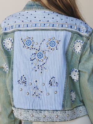 A close-up of the back of a light-wash denim jacket layered with a striped fabric panel, intricate floral embroidery, beading, sequins, and lace trim — the kind of piece that looks like it collected stories from everywhere it's been.