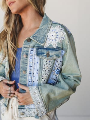 A close-up of a heavily embellished light-wash denim jacket layered over a blue ribbed tank — the kind of jacket that tells a story before you even say hello, with pearl beading, lace appliqué patches, floral embroidery, and striped fabric insets mak