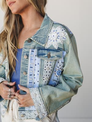 A close-up of a heavily embellished light-wash denim jacket layered over a blue ribbed tank — the kind of jacket that tells a story before you even say hello, with pearl beading, lace appliqué patches, floral embroidery, and striped fabric insets mak