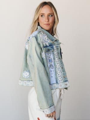 A light-wash denim jacket covered in floral appliqués, lace trim, pearl beading, and embroidery — the kind of piece that looks like it collected stories along the way — styled over white wide-leg pants with a soft studio backdrop.