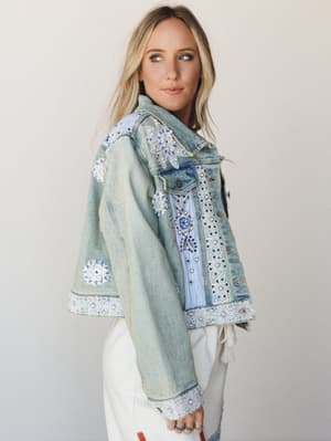 A light-wash denim jacket covered in floral appliqués, lace trim, pearl beading, and embroidery — the kind of piece that looks like it collected stories along the way — styled over white wide-leg pants with a soft studio backdrop.