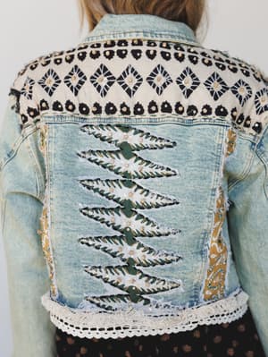 A close-up back view of a heavily embellished light wash denim jacket featuring mixed fabric patchwork, geometric block print across the yoke, vertical slash cutouts revealing a floral-printed underlayer, sequin and bead detailing, and a crochet lace