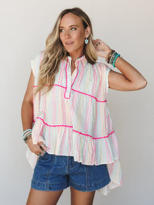 A breezy, tiered sleeveless top in ivory with multicolor stripes and hot pink stitch trim, styled with denim shorts and stacked bracelets — the kind of outfit that looks effortless at a farmers market or a beach-town lunch.