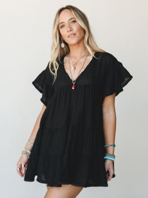 A breezy black mini dress with tiered ruffle layers and flutter sleeves, styled with stacked beaded bracelets and layered charm necklaces — the kind of effortless outfit that works just as hard at brunch as it does at a summer concert.
