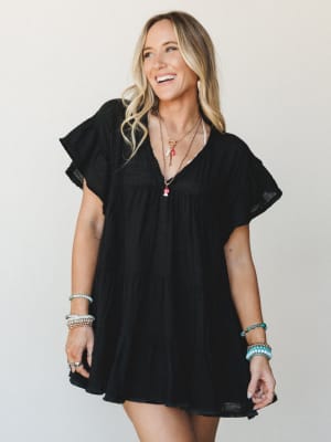 A breezy black mini dress with tiered skirt and flutter sleeves, styled with layered charm necklaces and stacked beaded bracelets — the kind of effortless outfit that works just as hard at brunch as it does at a summer concert.