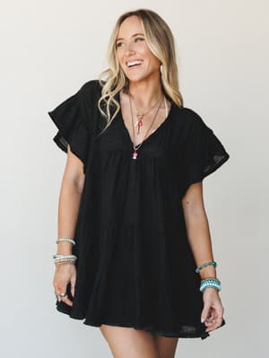 A breezy black mini dress with tiered skirt and flutter sleeves, styled with layered charm necklaces and stacked beaded bracelets — the kind of effortless outfit that works just as hard at brunch as it does at a summer concert.