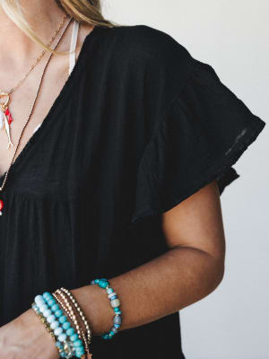 A close-up styled shot of a black flutter-sleeve dress layered with stacked turquoise and gold beaded bracelets and layered charm necklaces — the kind of outfit that looks effortless but absolutely has a personality.