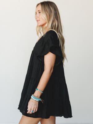 A breezy black tiered mini dress with flutter sleeves shot in profile — styled with stacked turquoise and gold bracelets that give it just enough boho personality to keep it from feeling too serious.