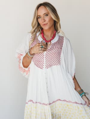A flowy white maxi dress with red embroidered geometric yoke detail, yellow floral hem tier, and button-front closure — styled with stacked bracelets, silver rings, and a knotted red bandana necklace for an effortless boho-summer feel.