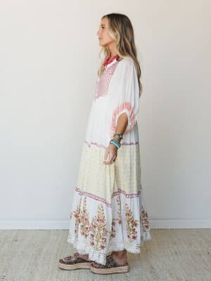 A flowy white maxi dress with mixed block print panels — floral hem border, small yellow ditsy print skirt tier, and red embroidered bib — styled with stacked turquoise bracelets, a red bandana neckerchief, and espadrille platform sandals for a free-