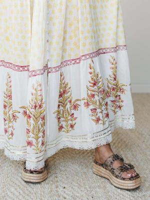A close-up of a flowing maxi dress hem in cream and yellow block print with a bold floral border, lace trim, and gold accent stripe — styled with leopard print platform espadrille sandals on a woven rug.
