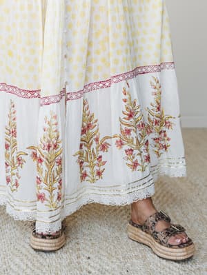 A close-up of a flowing maxi dress hem in cream and yellow block print with a bold floral border, lace trim, and gold accent stripe — styled with leopard print platform espadrille sandals on a woven rug.