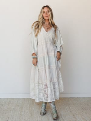 A flowy tiered maxi dress in soft sage and cream with wide lace panels throughout, styled with embroidered cowboy boots and layered boho jewelry — the kind of dress that makes you feel like you wandered into a garden party and never left.