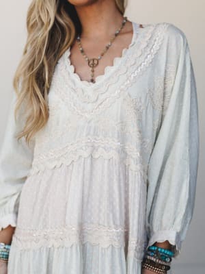 A dreamy close-up of a flowy maxi dress in soft cream and pale blue, covered in delicate sequin trim, scalloped lace bands, and beaded embroidery — styled with turquoise bracelets and a peace-sign pendant necklace for that effortless boho-romantic fe