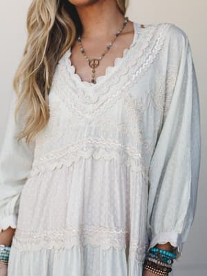 A dreamy close-up of a flowy maxi dress in soft cream and pale blue, covered in delicate sequin trim, scalloped lace bands, and beaded embroidery — styled with turquoise bracelets and a peace-sign pendant necklace for that effortless boho-romantic fe