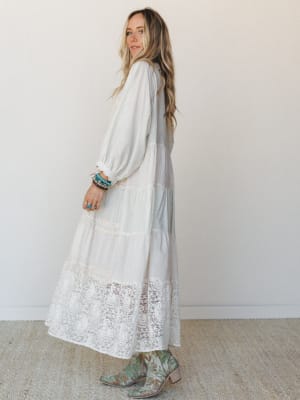 A cream lace-hem maxi dress with long bishop sleeves and tiered skirt panels, styled with turquoise western boots and stacked boho bracelets — the kind of outfit that could go from a backyard ceremony to a Sunday farmer's market without missing a bea