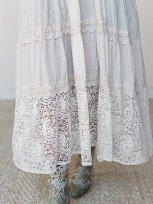 A close-up detail shot of a tiered maxi dress hem in soft dusty blue with Swiss dot texture, lace trim bands, and a sheer paisley lace hem panel — styled with embroidered cowboy boots for that effortless boho-meets-western feel.