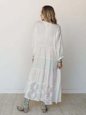 Shot from the back, a cream tiered maxi dress with a lace hem panel is styled with embroidered cowboy boots and turquoise stacked bracelets — romantic and a little Western, in the best way.
