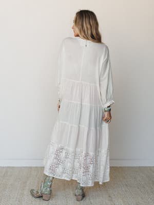 Shot from the back, a cream tiered maxi dress with a lace hem panel is styled with embroidered cowboy boots and turquoise stacked bracelets — romantic and a little Western, in the best way.