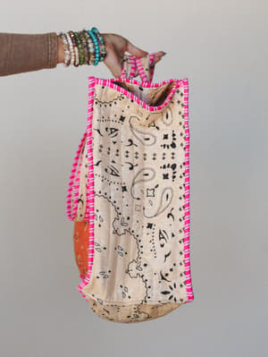 A stacked-bracelet hand holds up a bandana-print tote with hot pink whipstitch edging — the kind of bag that looks like it was made from a favorite vintage scarf and goes everywhere without trying.