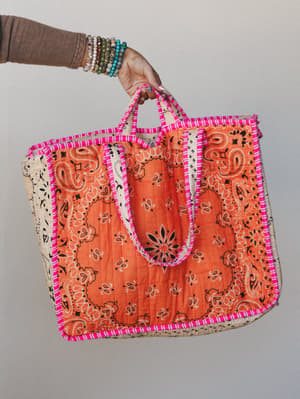 A quilted bandana-print tote in bold orange and cream is held up by a hand stacked with beaded bracelets — the hot pink whipstitch trim ties it all together into something that feels equal parts nostalgic and playful.