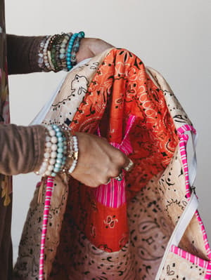 A close-up of a model opening a roomy patchwork tote bag — bandana-print panels in orange, cream, and hot pink with white canvas handles, held by bracelet-stacked hands that make the whole thing feel like a mood.