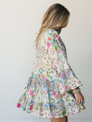 A flowy mini dress in a vibrant mixed floral print gets the full spin treatment — tiered ruffled hem, bell sleeves, and stacked boho jewelry making it feel like something you'd reach for every warm-weather occasion without overthinking it.