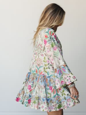 A flowy mini dress in a vibrant mixed floral print gets the full spin treatment — tiered ruffled hem, bell sleeves, and stacked boho jewelry making it feel like something you'd reach for every warm-weather occasion without overthinking it.