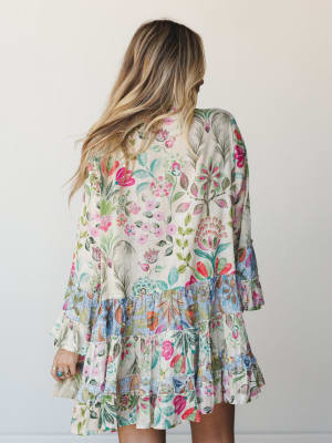 A back-facing shot of a flowy mini dress in a vibrant mixed floral print — cream base with a contrasting blue floral ruffle hem and bell sleeves, styled with stacked rings and relaxed waves for an effortlessly garden-party feel.