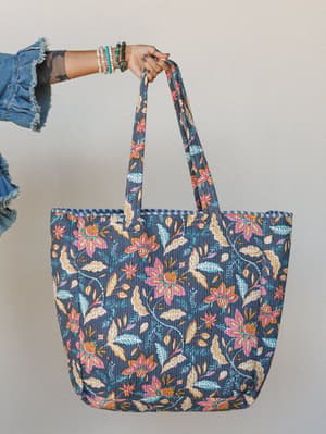 A tattooed hand stacked with beaded bracelets holds up a large quilted tote covered in a painterly floral vine print — the kind of bag that goes from farmers market to weekend getaway without missing a beat.