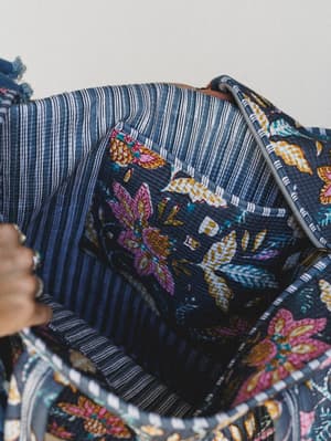 A close-up interior shot of a quilted tote bag held open by a ringed hand, revealing a bold botanical floral lining against a navy striped interior — the kind of bag that surprises you every time you reach inside.