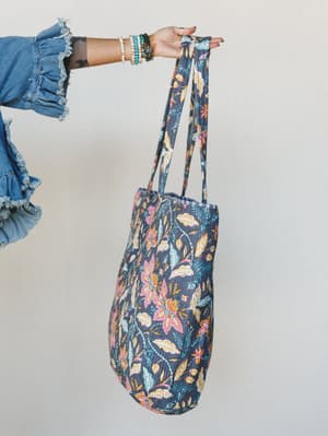 A tattooed arm draped in stacked bracelets holds up a quilted floral tote bag against a soft neutral wall — the kind of bag that makes a farmers market run feel like a whole moment.