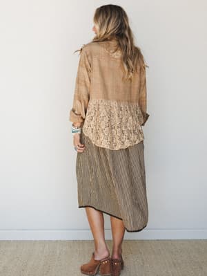 Shot from the back, a model in a warmly layered boho outfit — a woven mocha jacket with a floral lace panel at the hem, worn over a black-and-tan striped midi skirt, finished with wood-platform clogs and a stack of turquoise and cream bracelets.