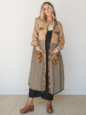 A long patchwork duster coat in warm mocha and tan tones layered over dark denim, styled with wood-studded clogs and stacked bracelets — earthy, collected, and the kind of piece that makes a whole outfit feel intentional.