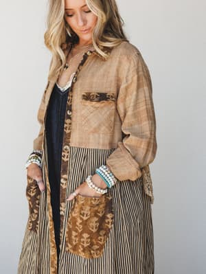 A long patchwork duster jacket in warm mocha tones — mixing block-print florals, thin stripes, and woven textures — layered over a dark denim dress with lace cami peeking through, styled with stacked beaded bracelets and a gold chain necklace.