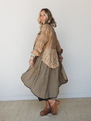 A flowy, patchwork-style maxi duster jacket in warm mocha tones spins mid-movement, revealing a striped skirt underneath and lace panel detail — the kind of piece that looks like it has a whole backstory.