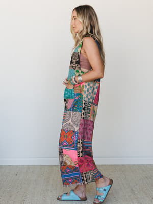 A side-profile shot of a model in a bold, patchwork-print wide-leg jumpsuit layered over a mauve bralette, styled with light blue buckle sandals and stacked bracelets — the kind of outfit that has its own personality before you even walk in.