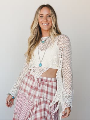 A sun-kissed blonde model smiles warmly in a cream open-knit crochet poncho layered over a blush plaid tiered skirt, styled with turquoise jewelry for a free-spirited, effortlessly boho feel.